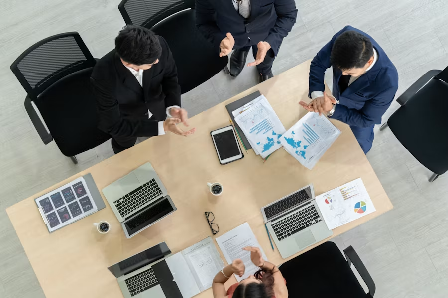 Four colleagues in business attire discuss documents and charts around a light wooden meeting table, seen from above.
