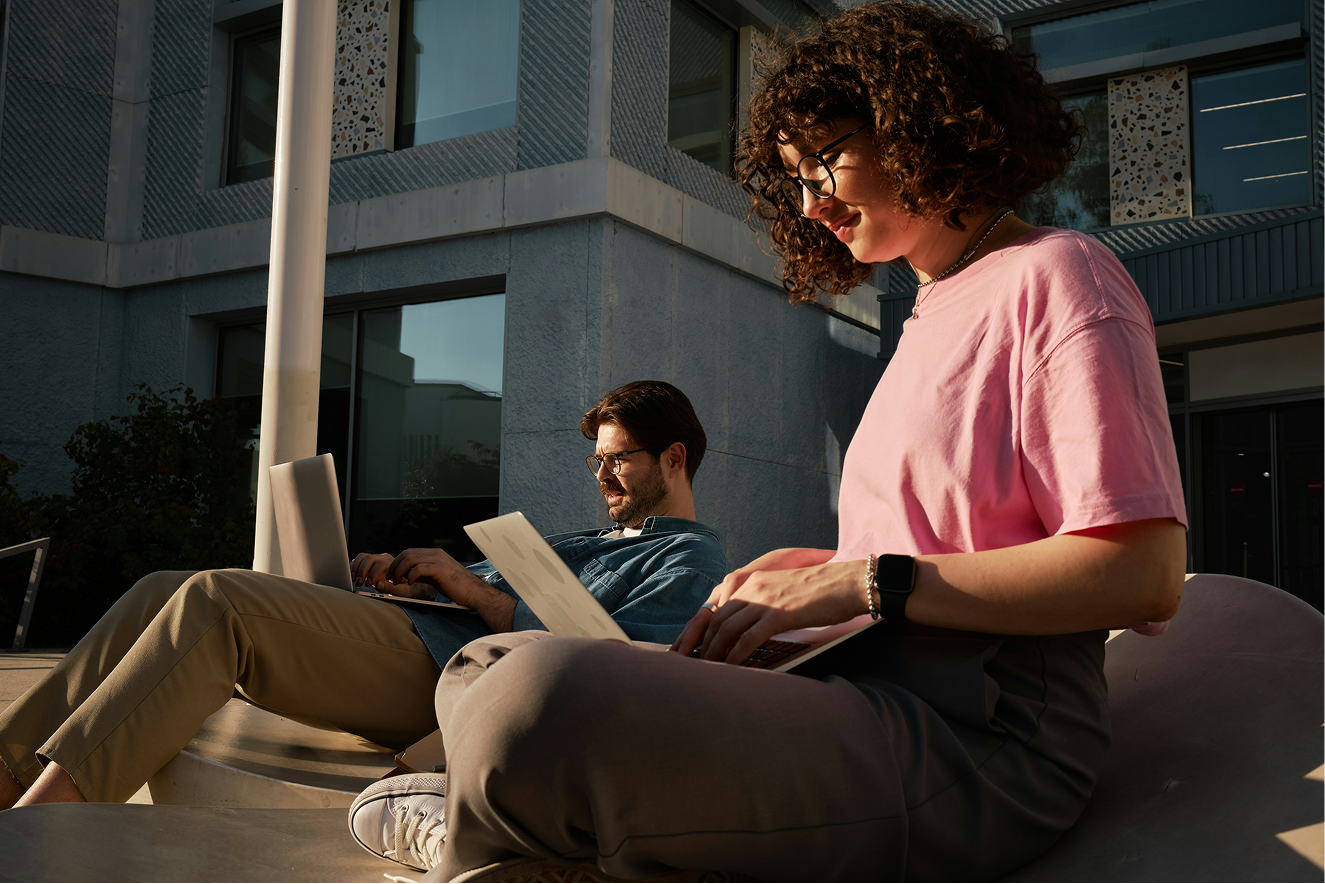 Two people working on laptops on concrete steps in warm afternoon light outside a modern building.