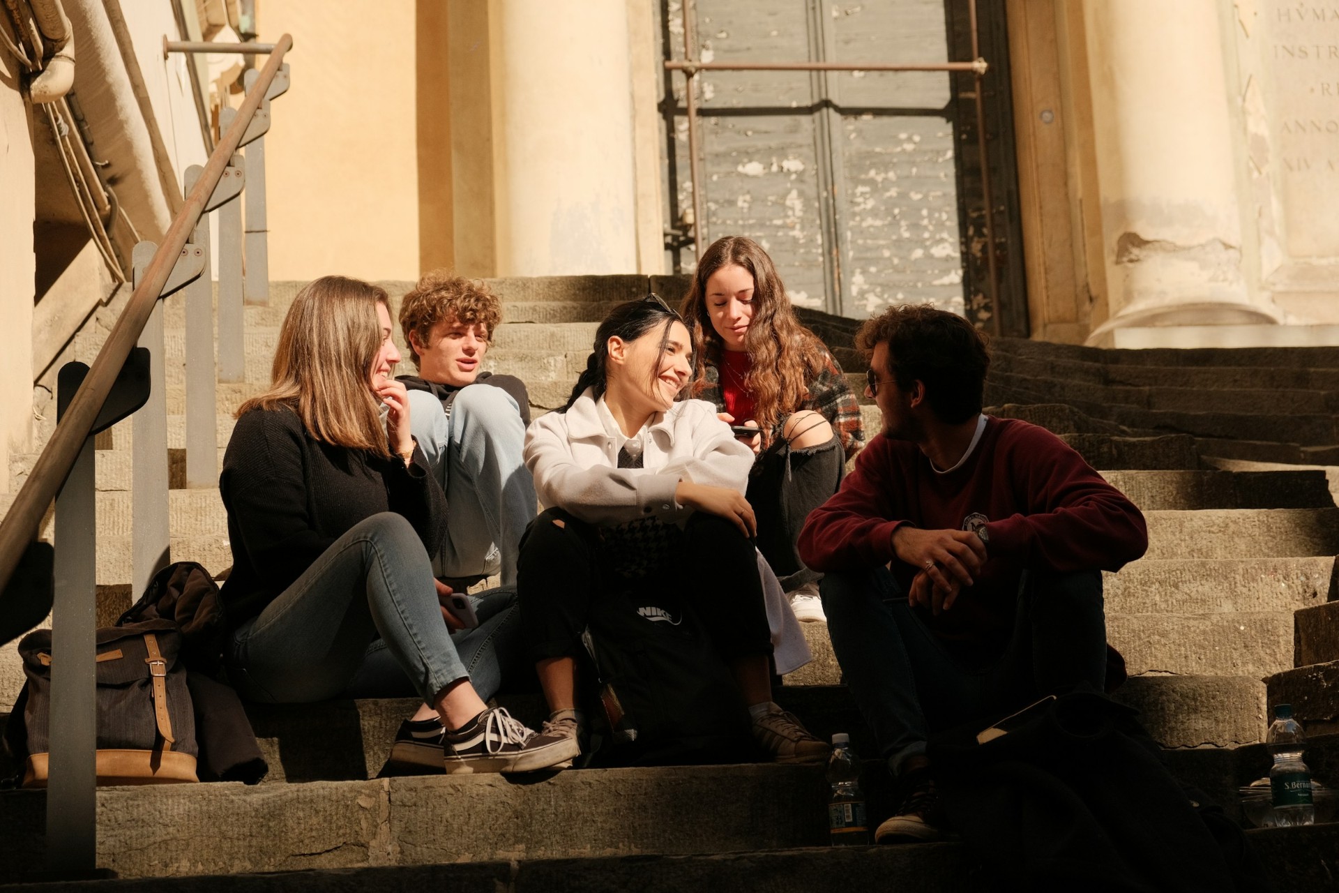 Five friends sitting and chatting on stone steps outside a historic building.