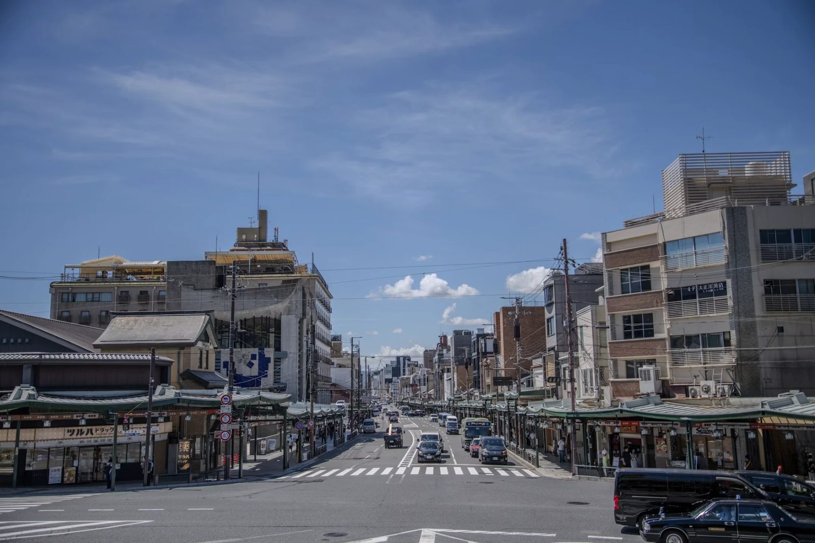 A wide Japanese city street on a clear day, lined with shopfront canopies, overhead cables and traffic heading into the distance.